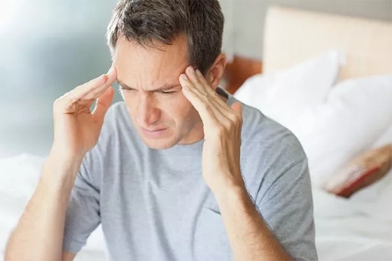 Man holding his head in discomfort, seated on a bed.