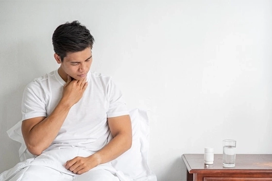 Man sitting on a bed holding his throat, appearing to experience discomfort, with a glass of water and medication on the bedside table.